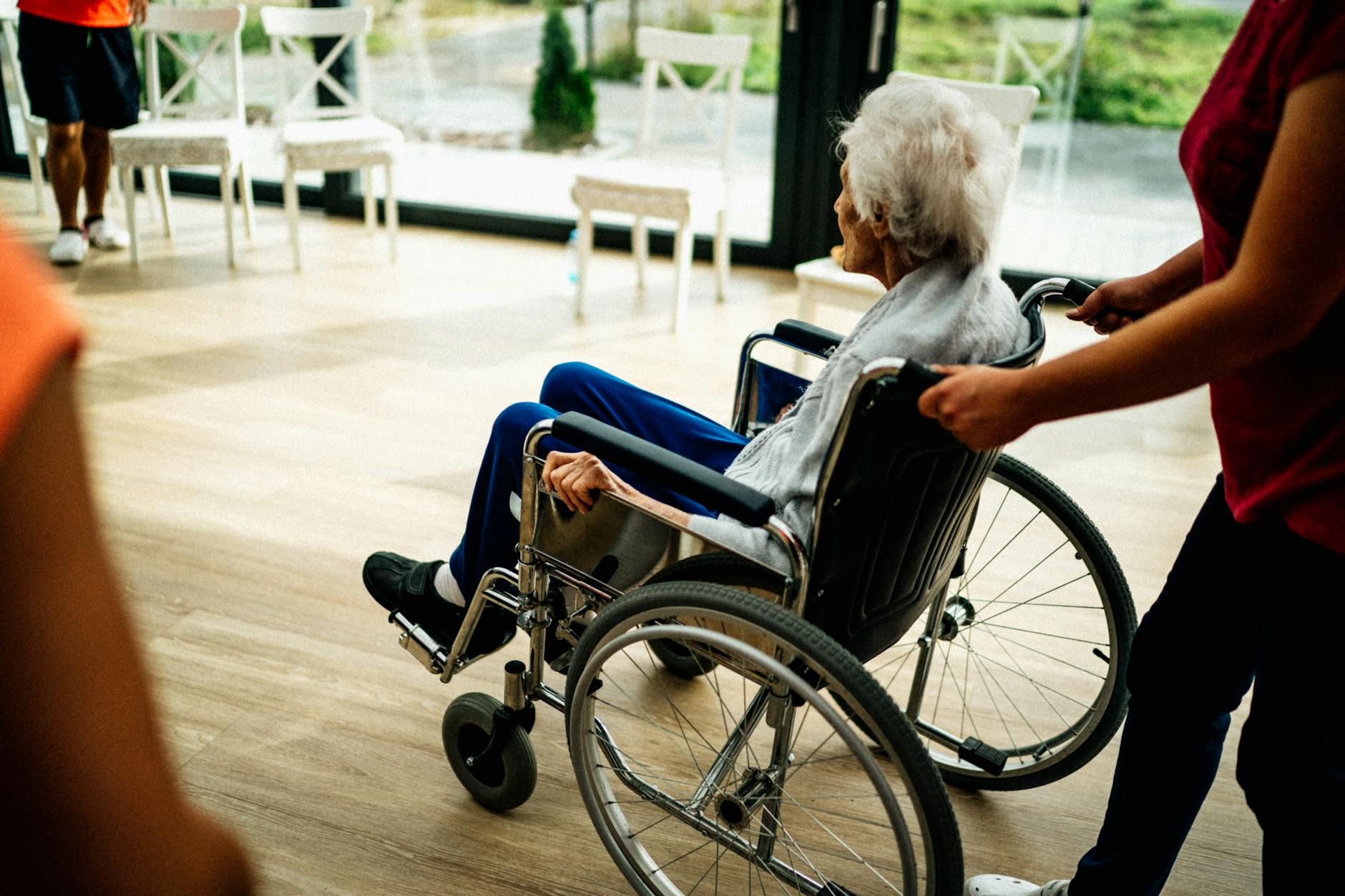 An elderly woman in a wheelchair being assisted by a caregiver indoors.