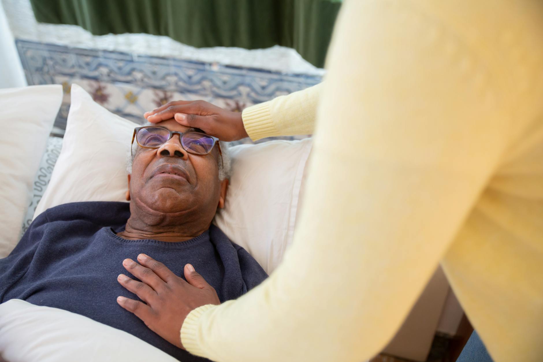 A caregiver checking the forehead of an elderly man lying in bed, providing health care and support.